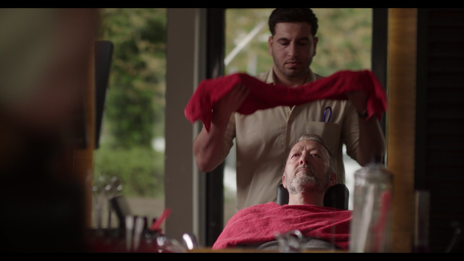 A barber draws a red towel over a reclined customer, a graded frame finished in post-production